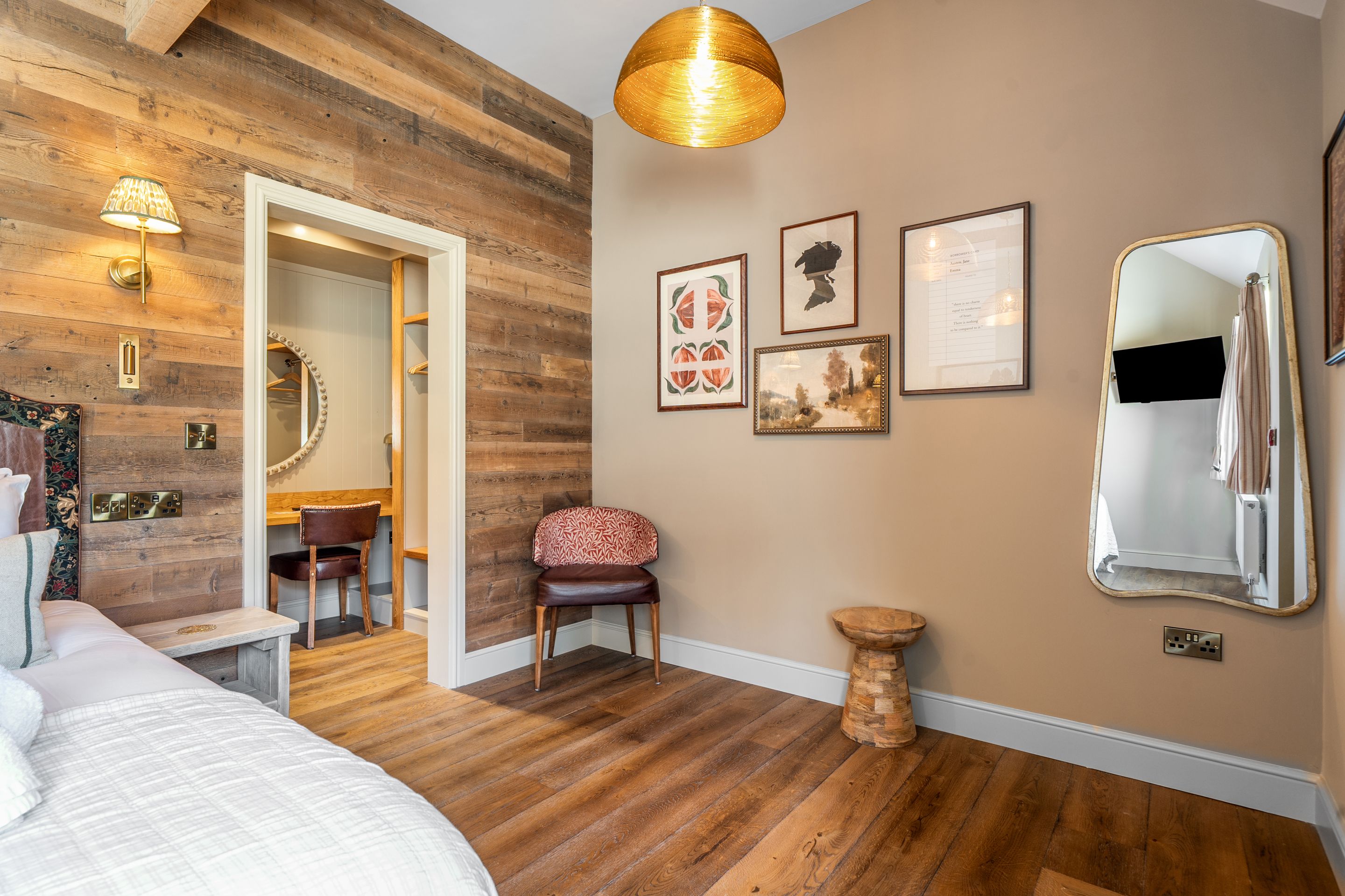Une pièce avec un plancher en bois, un lit, une chaise, une petite table, un miroir et des œuvres d’art accrochées au mur.