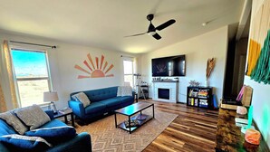 A living room with a blue sofa, a coffee table, a fireplace, a TV, and a ceiling fan. - Spectacular panoramic views on nearly 10 acres, Stargazing, Spa, Family Friendly (Twentynine Palms)