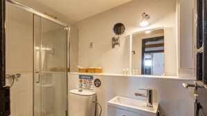 Uma casa de banho com cabine de chuveiro em vidro, sanita, lavatório e espelho. Há um aviso na parede a indicar que não se deve deitar lixo ou papel na sanita.