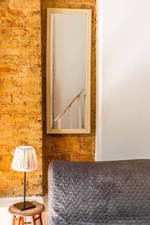 A room with a brick wall, a bed with a grey quilt, a lamp on a small table, and a mirror on the wall.