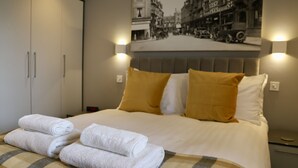 A neatly made bed with white and mustard pillows, a headboard with wall-mounted lights, and a folded white towel on the bed. - Apartment Seven (Shrewsbury)