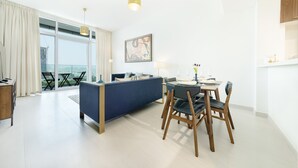 A modern living room with a blue sofa, wooden dining table, and chairs. There is a large window with curtains, a painting on the wall, and a view of a building outside. - Maison Privee - Superb 1BR apartment overlooking Zabeel Park and Dubai Frame (Dubai)