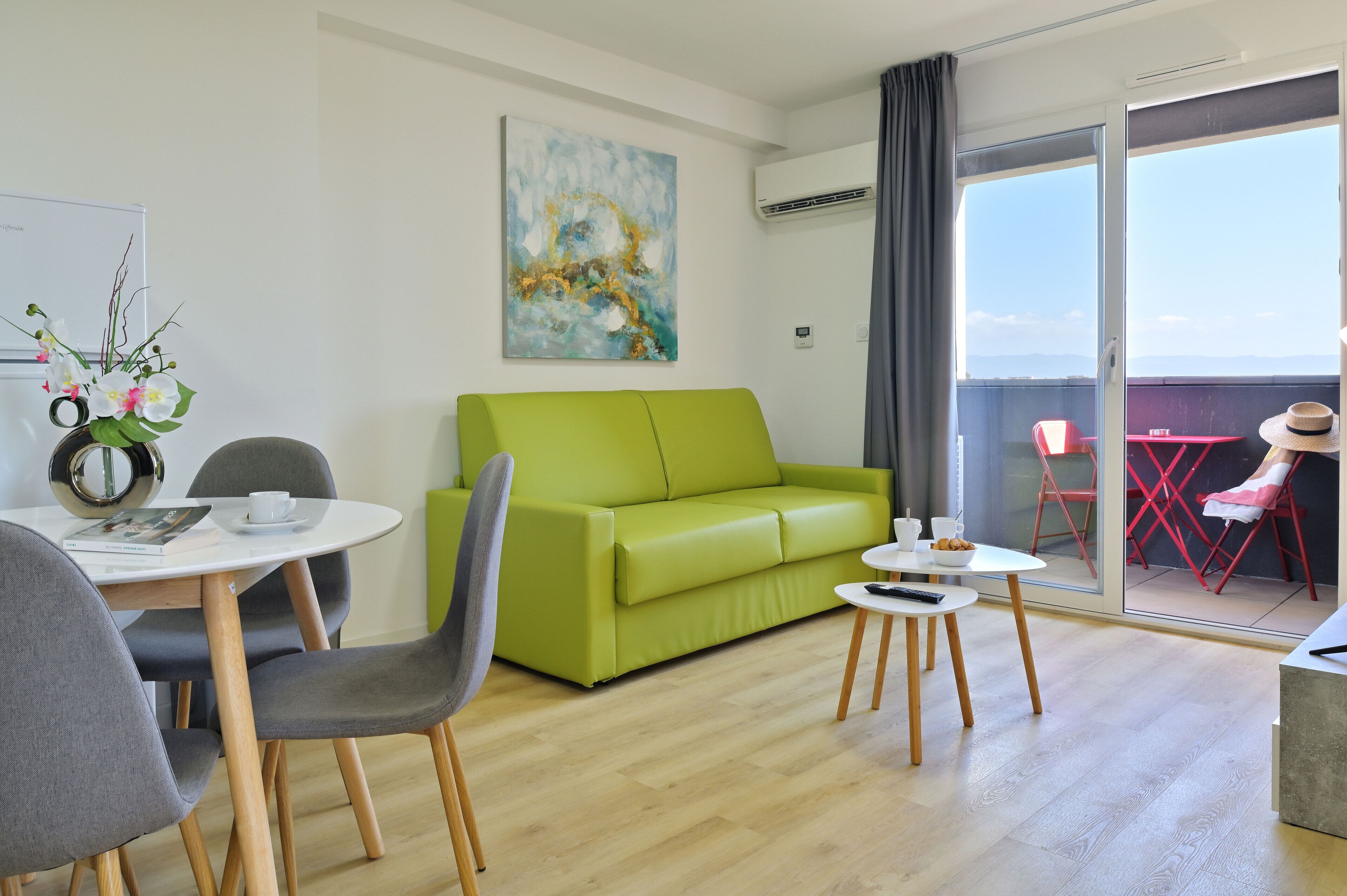 A modern living room with a green sofa, a dining table with chairs, a small side table, and a balcony with red chairs.