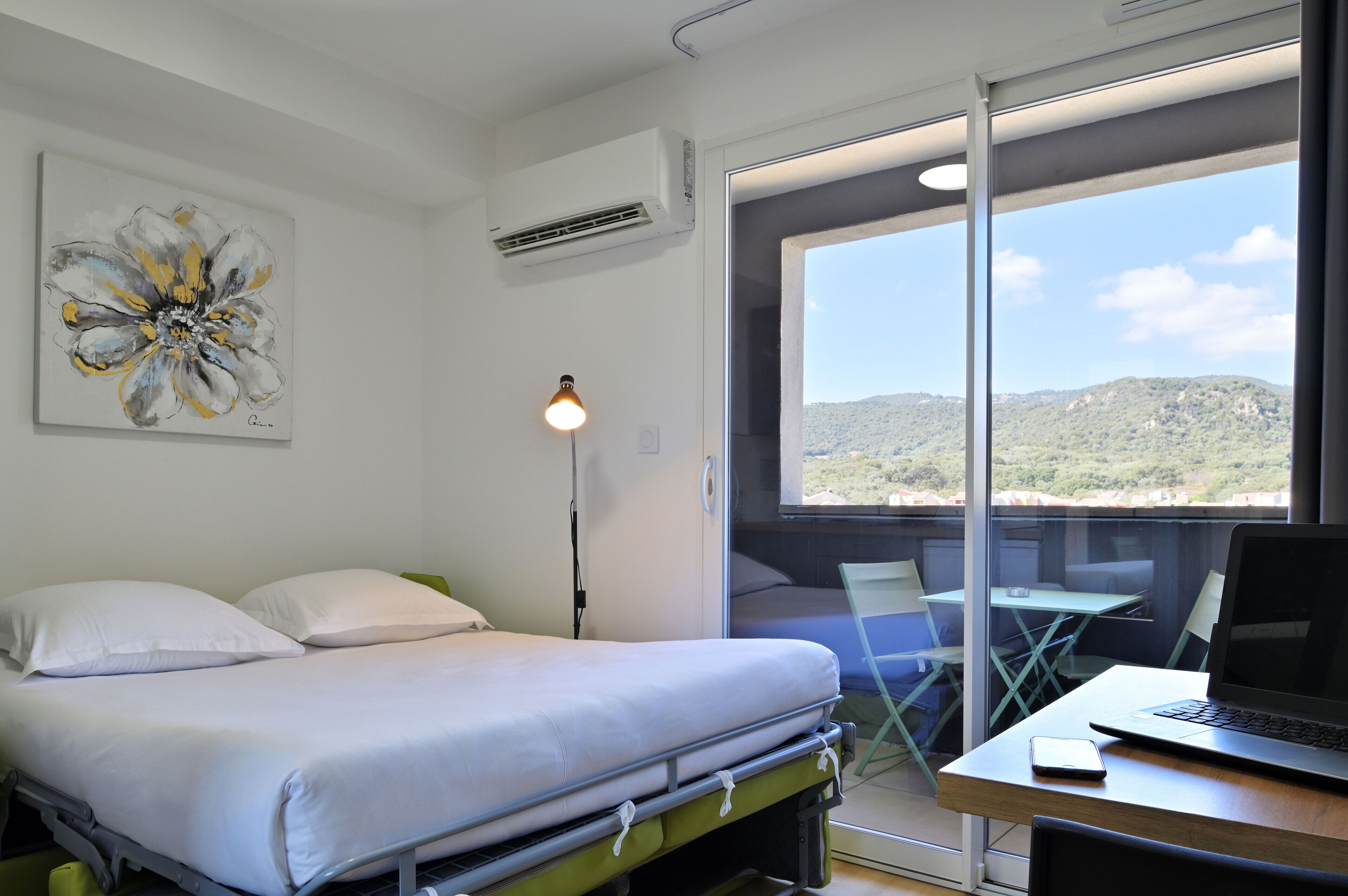 A room with a bed, a desk with a laptop, a chair, and a table. There is a window with a view of hills.