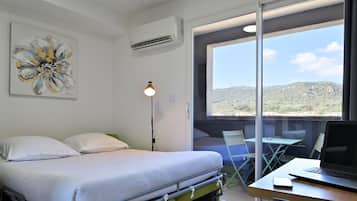 A room with a bed, a desk with a laptop, a chair, and a table. There is a window with a view of hills.