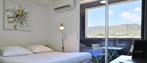 Quarto com cama, escrivaninha com laptop, cadeira e mesa. Janela com vista para colinas.