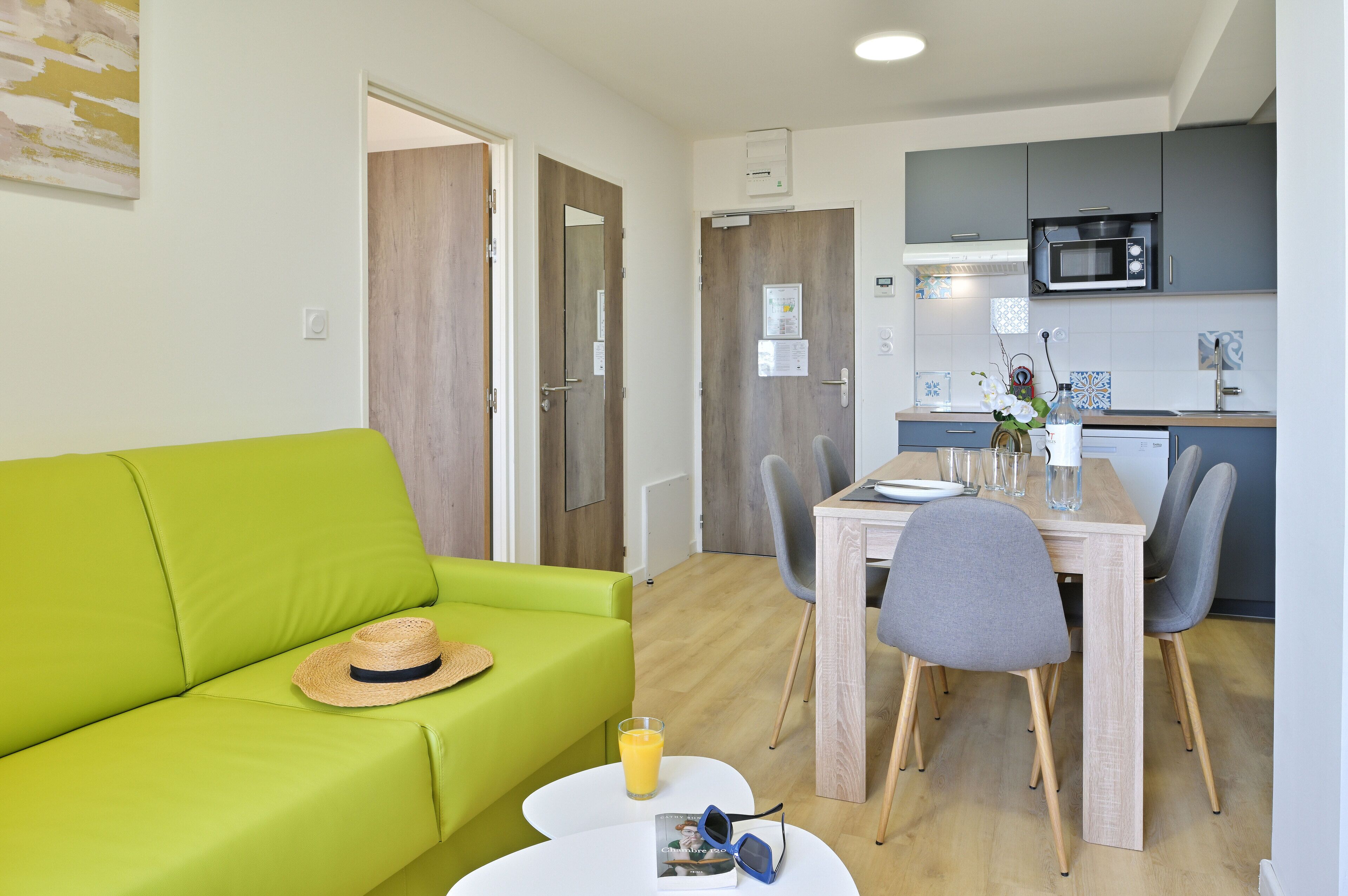A bright living room with a green sofa, a dining table with chairs, and a kitchen area with a microwave.