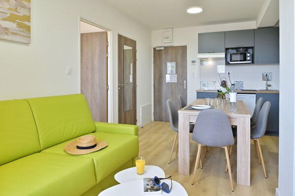 Un salón luminoso con un sofá verde, una mesa de comedor con sillas y una zona de cocina con microondas.
