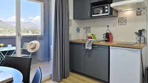 Une cuisine compacte avec des meubles gris, un four à micro-ondes et un petit réfrigérateur. On y trouve un coin repas avec une table et des chaises, et une vue sur l’extérieur grâce à une grande fenêtre.