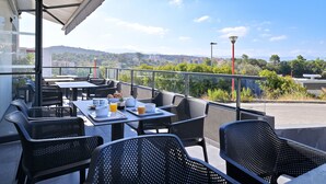 Un espace de restauration en plein air avec des tables et des chaises, une vue dégagée sur le paysage urbain et une partie abritée avec un mur de verre.