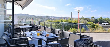 Un área de comedor al aire libre con mesas y sillas, vistas despejadas del paisaje urbano, y una sección cubierta con un muro de vidrio.