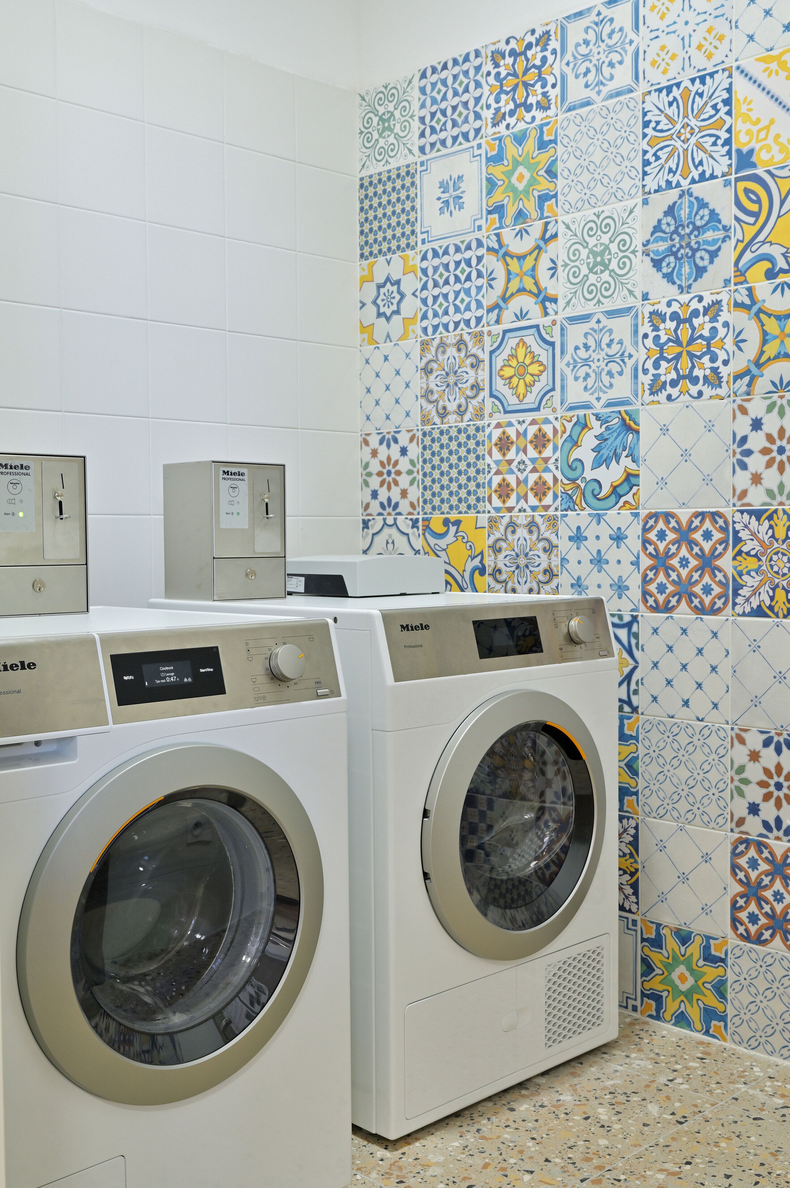 A laundry area with two washing machines and a control panel, set against a wall with patterned tiles.