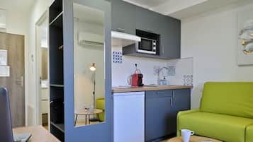 Cocina compacta con microondas, refrigerador pequeño y pileta. Hay un sofá verde y una mesa con una taza y un libro.