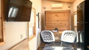 Un cuarto acogedor con techo de madera, un ventanal grande, dos sillones tapizados, una mesita con una planta y un refrigerador.