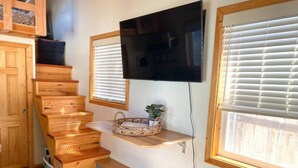 Una escalera de madera lleva a una zona de cocina con un televisor montado sobre una estantería, una ventana con persianas y una cesta en la estantería.