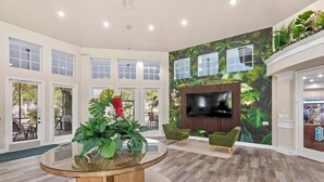 Un hall espacioso con una mesa de vidrio redonda, sillas verdes, un televisor grande y un mural tropical.