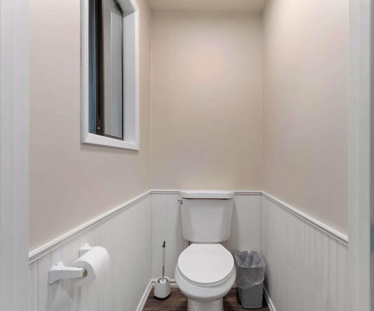A small, narrow bathroom with a toilet, a window, and a trash bin.