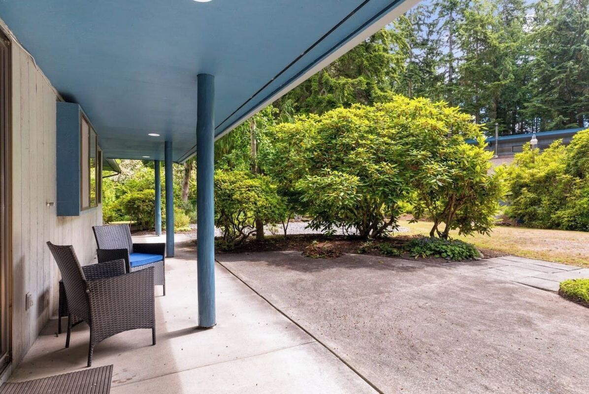 A covered patio with two wicker chairs and a table, surrounded by greenery and a building.