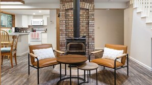 A cozy living room with a brick fireplace, two leather chairs, a round wooden table, and a kitchen in the background.