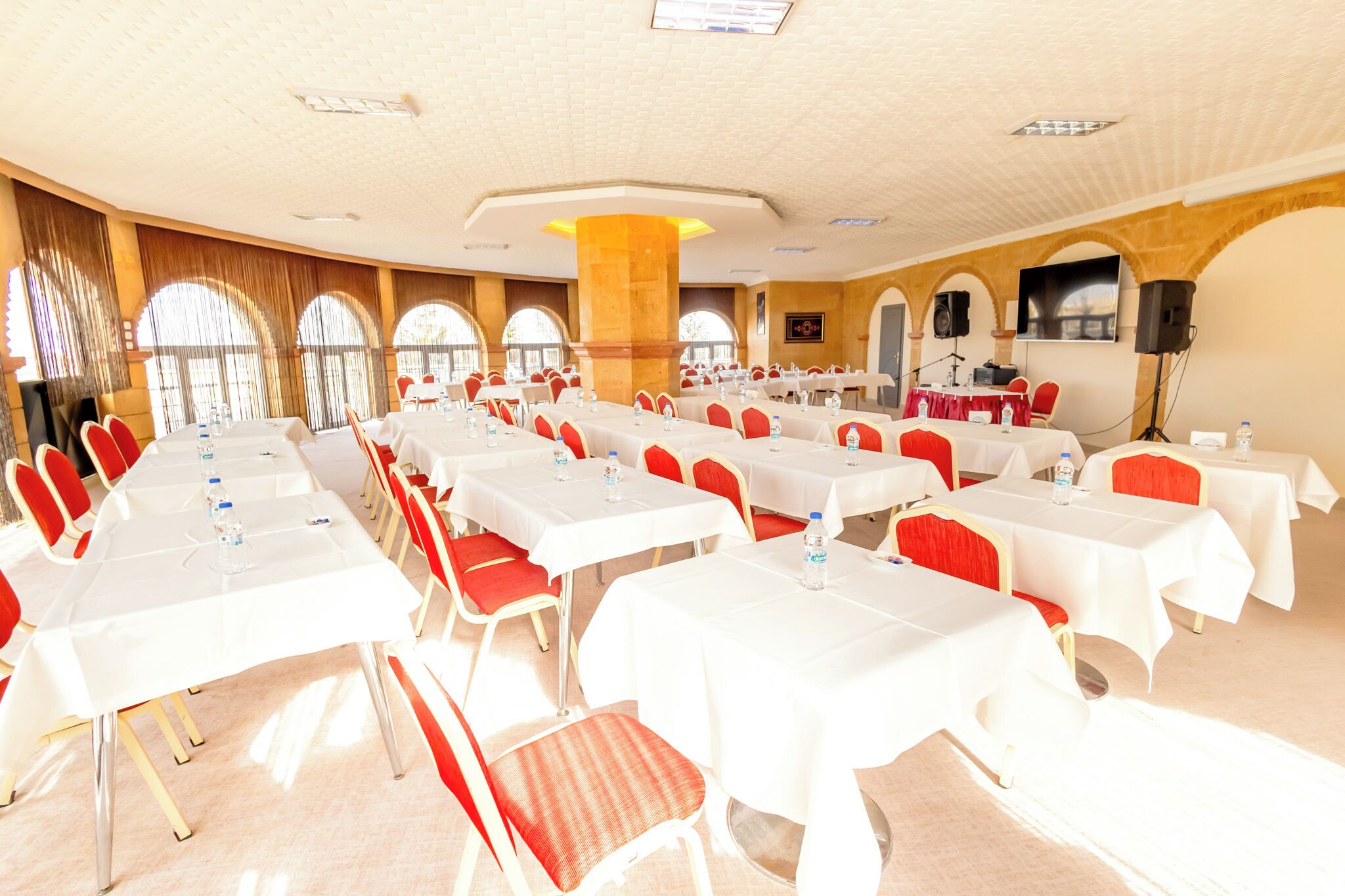 A conference room with multiple tables and chairs, large windows, and a stage with a screen and speakers.