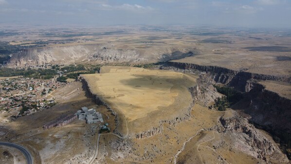 An aerial view of a rural landscape with a large open field, a small settlement with houses, and a winding road. - Continent Hotel Kapadokus Thermal (Guzelyurt)
