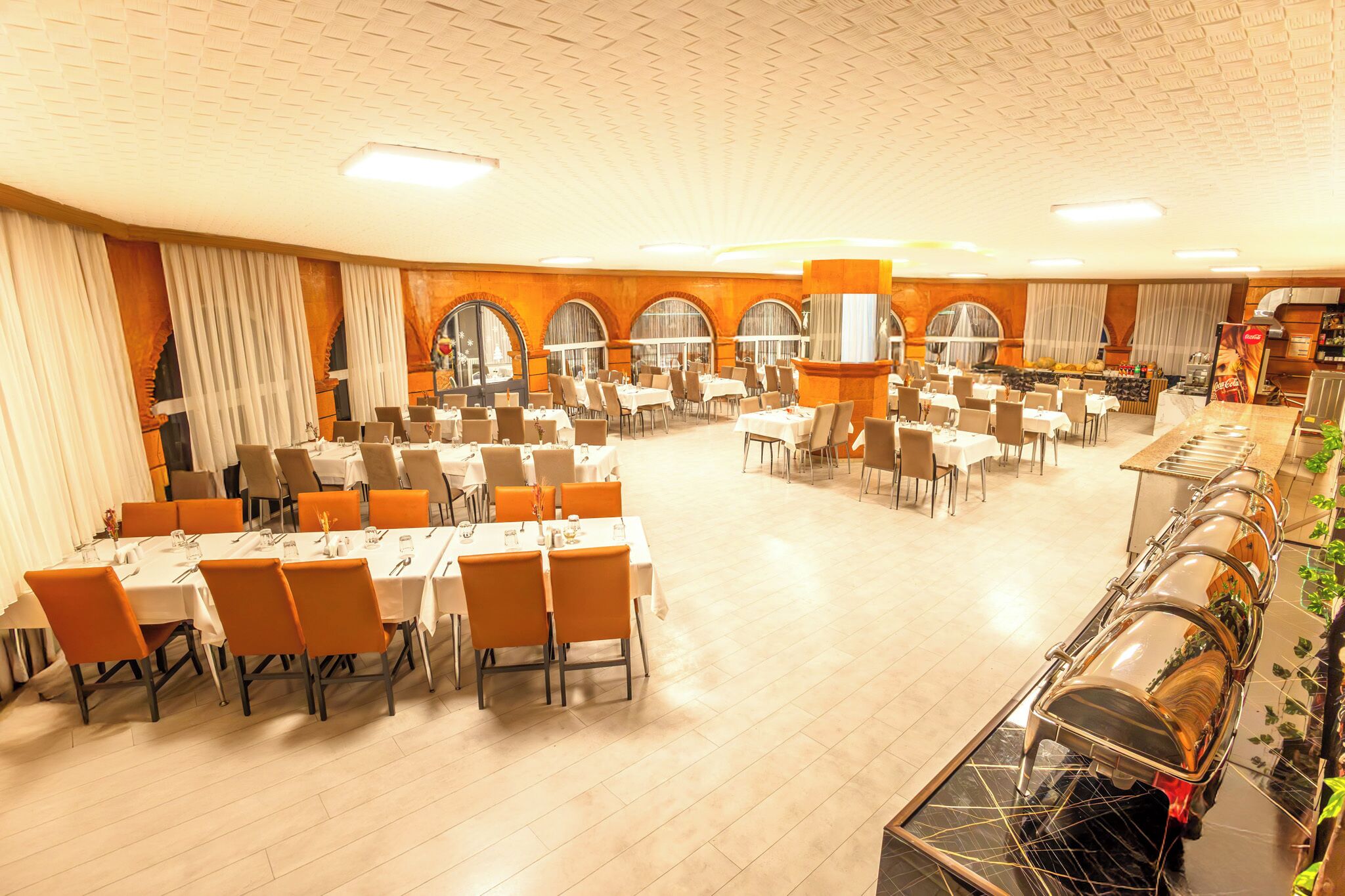 A spacious banquet hall with multiple tables and chairs, large windows with curtains, and a central column.