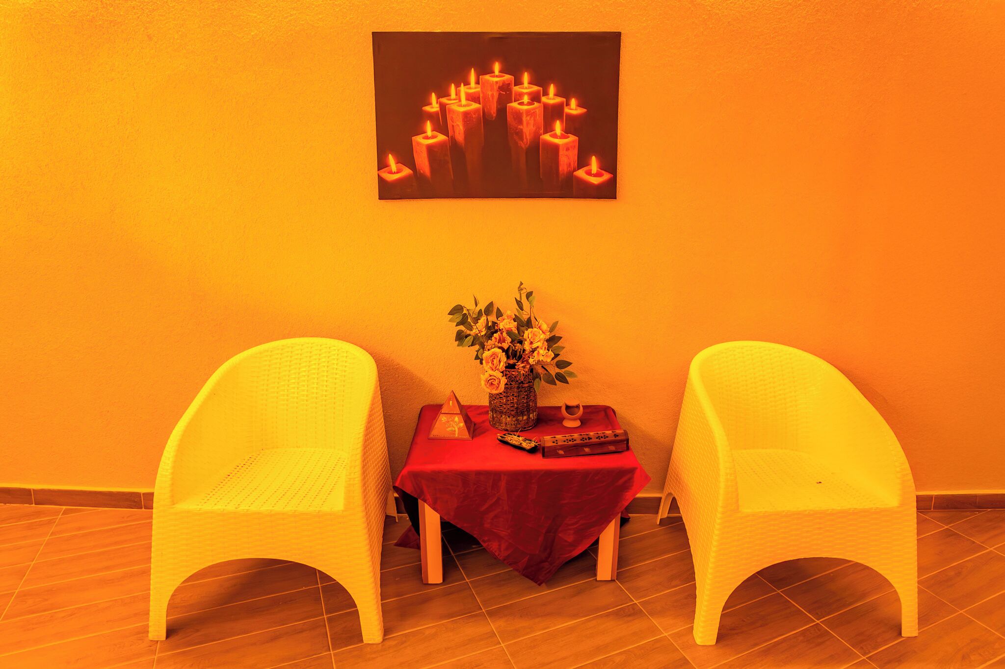 Una habitación con dos sillas blancas, una mesita con mantel rojo, un florero con flores y un cuadro de velas en la pared.