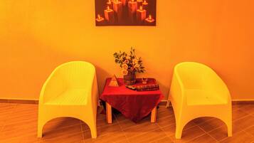 Una habitación con dos sillas blancas, una mesita con mantel rojo, un florero con flores y un cuadro de velas en la pared.