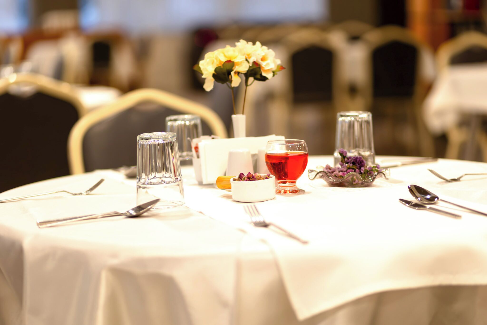 A dining table set with cutlery, glasses, a vase with flowers, and a glass of red liquid.