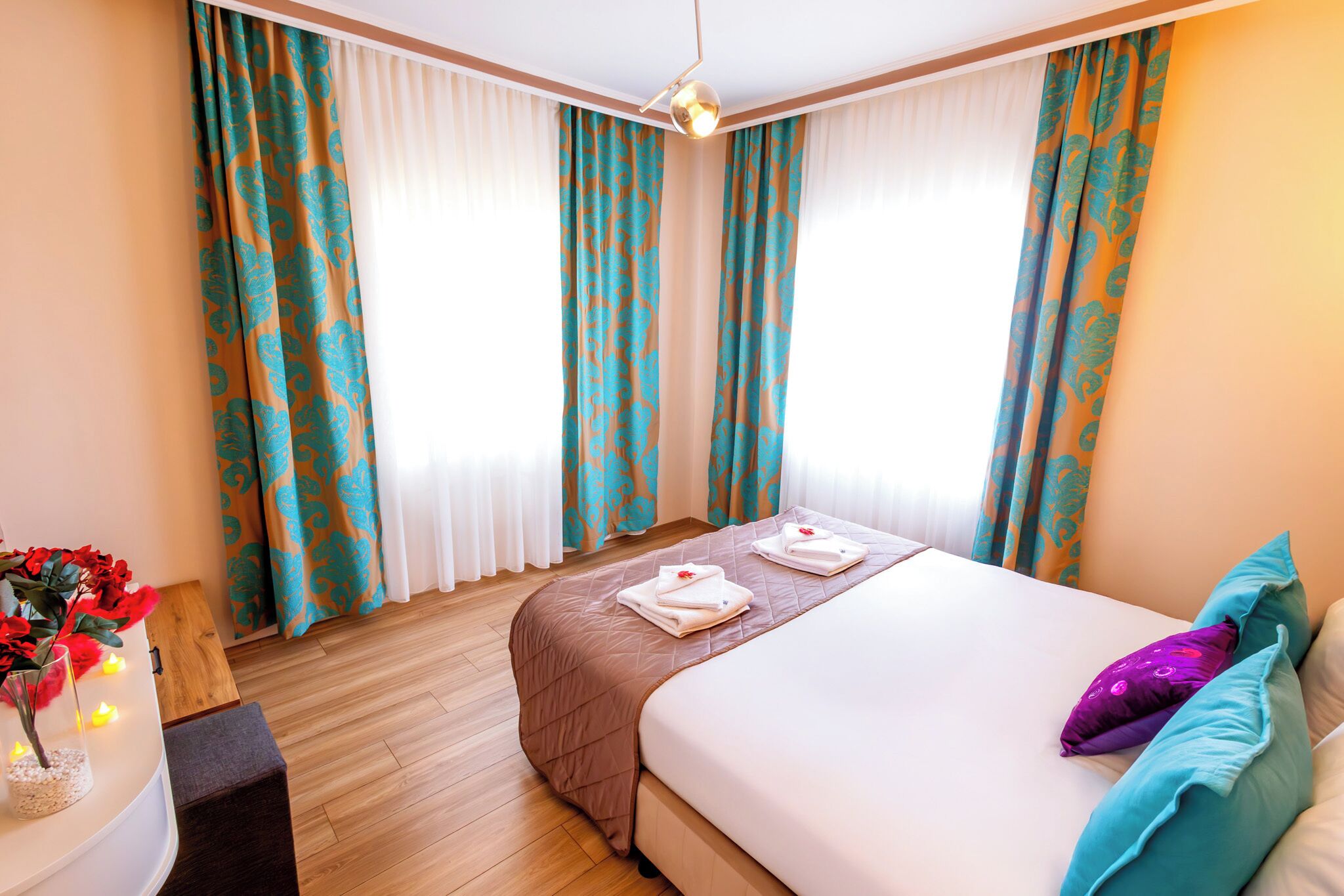 Quarto de hotel com uma cama, duas toalhas, um vaso com flores e uma escrivaninha com abajur.