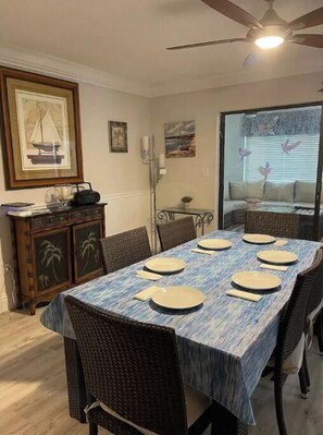Uma sala de jantar com mesa posta para seis pessoas, um ventilador de teto e vista para o exterior através de uma janela.