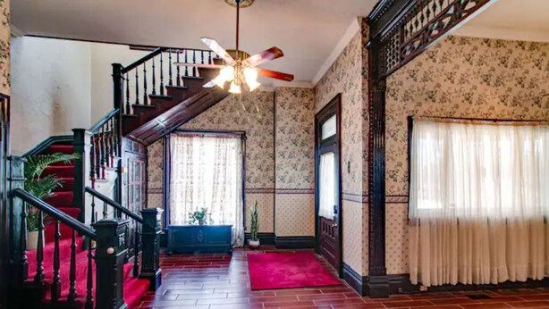A staircase with a wooden handrail and a red carpet leading to a room with floral wallpaper and a ceiling fan.