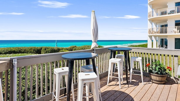 Una terraza con mesas y sillas que da a una playa, una sombrilla blanca y un edificio con balcones.