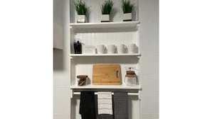 Eine weiße, geflieste Wand mit drei Regalen. Das oberste Regal beherbergt drei kleine Blumentöpfe. In der mittleren Ablage befinden sich eine Kaffeemaschine, ein Holzschneidebrett und ein Glas. Das untere Regal zeigt drei hängende Handtücher.