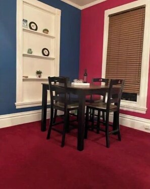 A dining area with a round table and four chairs, a white shelving unit with decorative items, and a window with blinds.