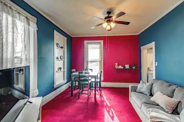 A room with a red carpet, a ceiling fan, a dining table with chairs, a grey sofa, and a window with blinds.