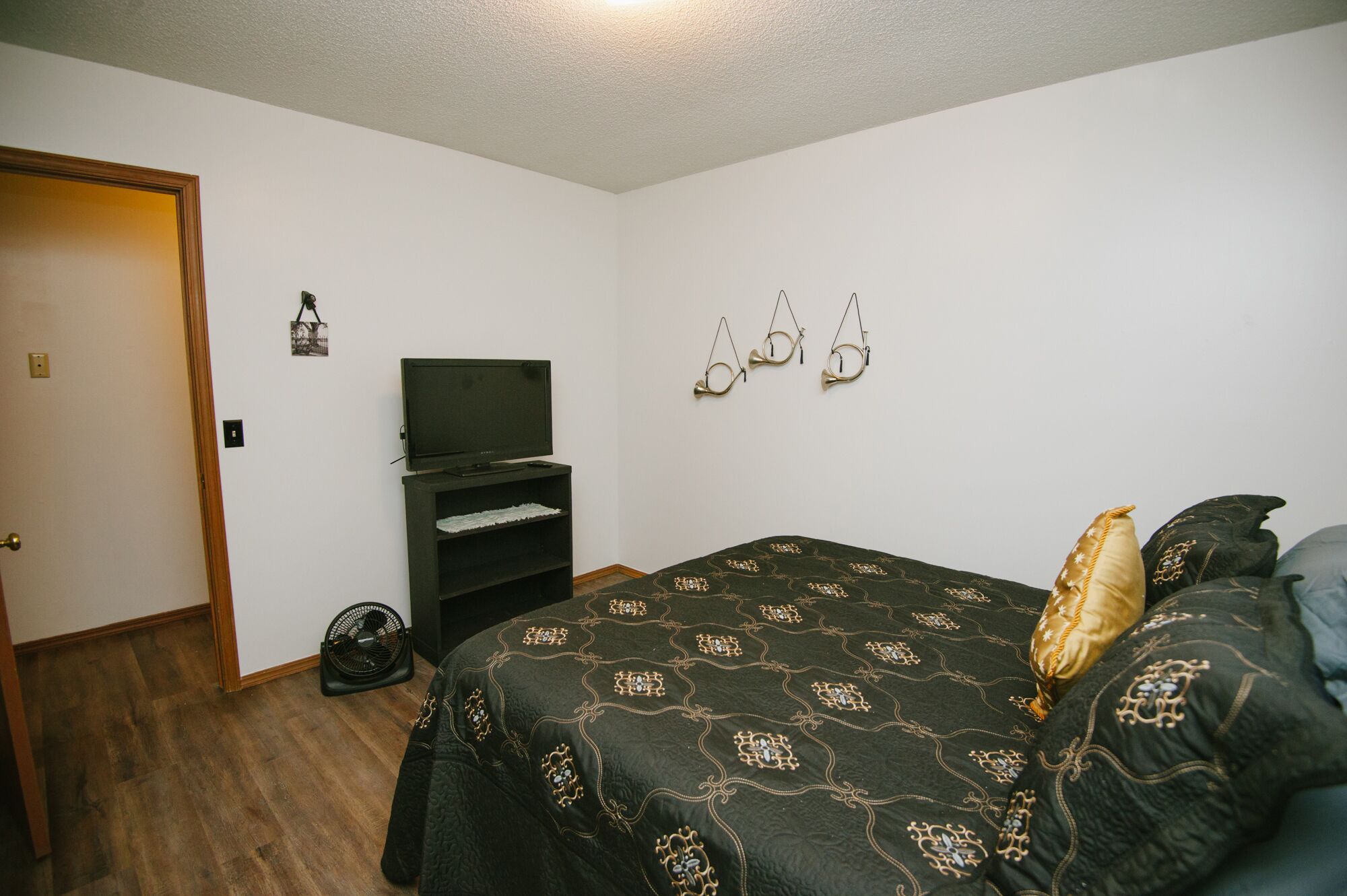 Un dormitorio con cama, televisor sobre un mueble, ventilador y una puerta.