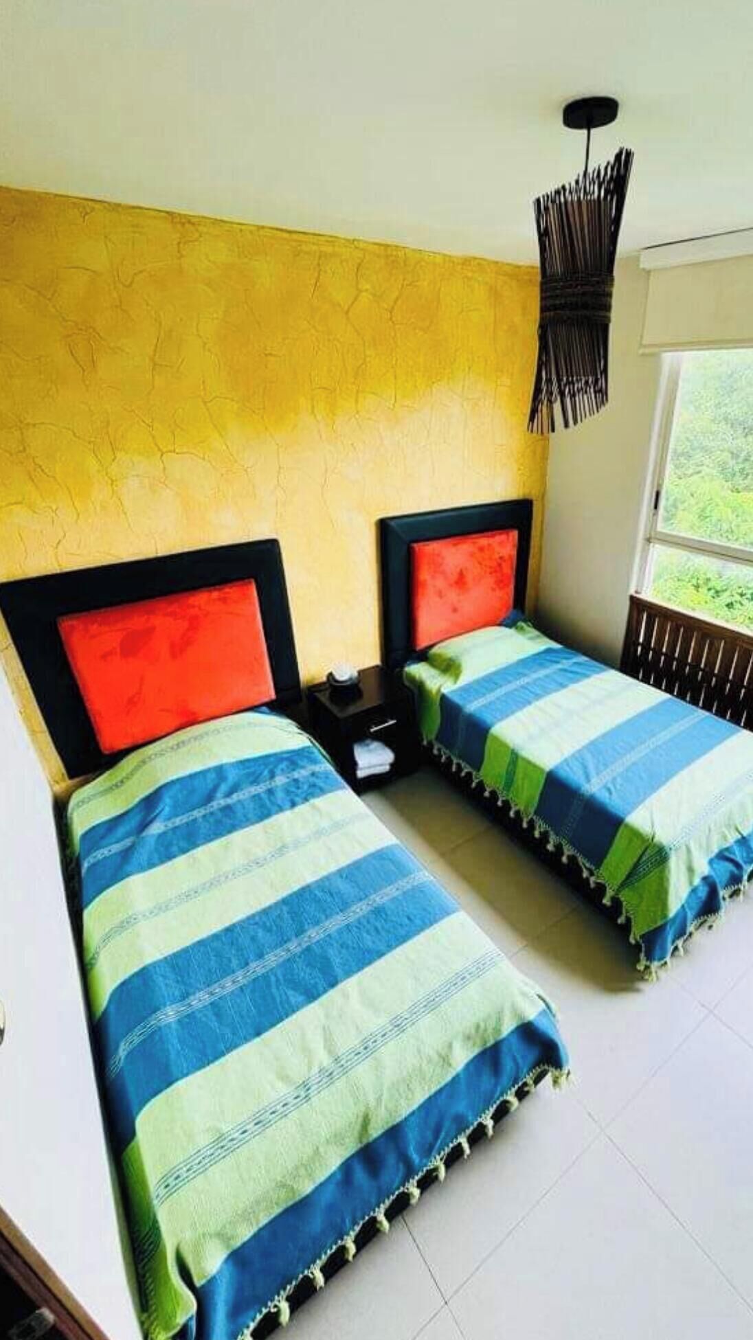 Una habitación con dos camas, cada una con una colcha a rayas y una almohada roja. Hay una mesita de noche con una lámpara entre las camas.