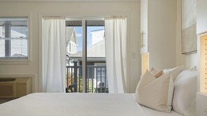 A bedroom with a large bed, white curtains, a view of a balcony, and a window with blinds. - Whale's Tale - Boutique Studio Condo w/Pool 🐳 (Hampton)