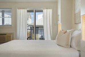 A bedroom with a large bed, white curtains, a view of a balcony, and a window with blinds.
