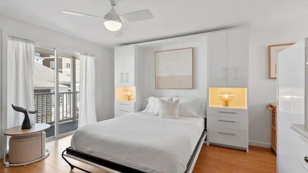 A modern hotel room with a bed, bedside tables, a ceiling fan, and a view of the outside balcony. - Whale's Tale - Boutique Studio Condo w/Pool 🐳 (Hampton)