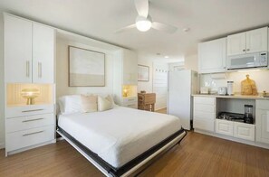 A bedroom with a bed, bedside tables, a desk, and a kitchenette with a microwave and a blender. - Whale's Tale - Boutique Studio Condo w/Pool 🐳 (Hampton)