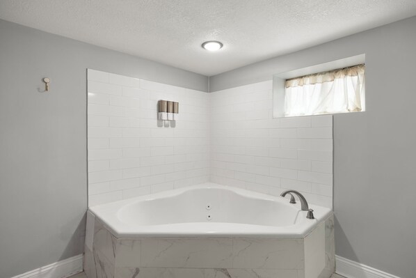 A corner bathroom with a white bathtub, a window with a curtain, and a wall-mounted light fixture.