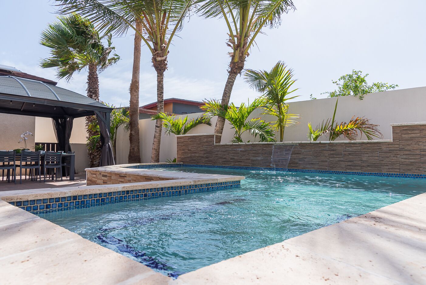 A swimming pool with tiled edges, surrounded by palm trees and a patio area with chairs and a table.
