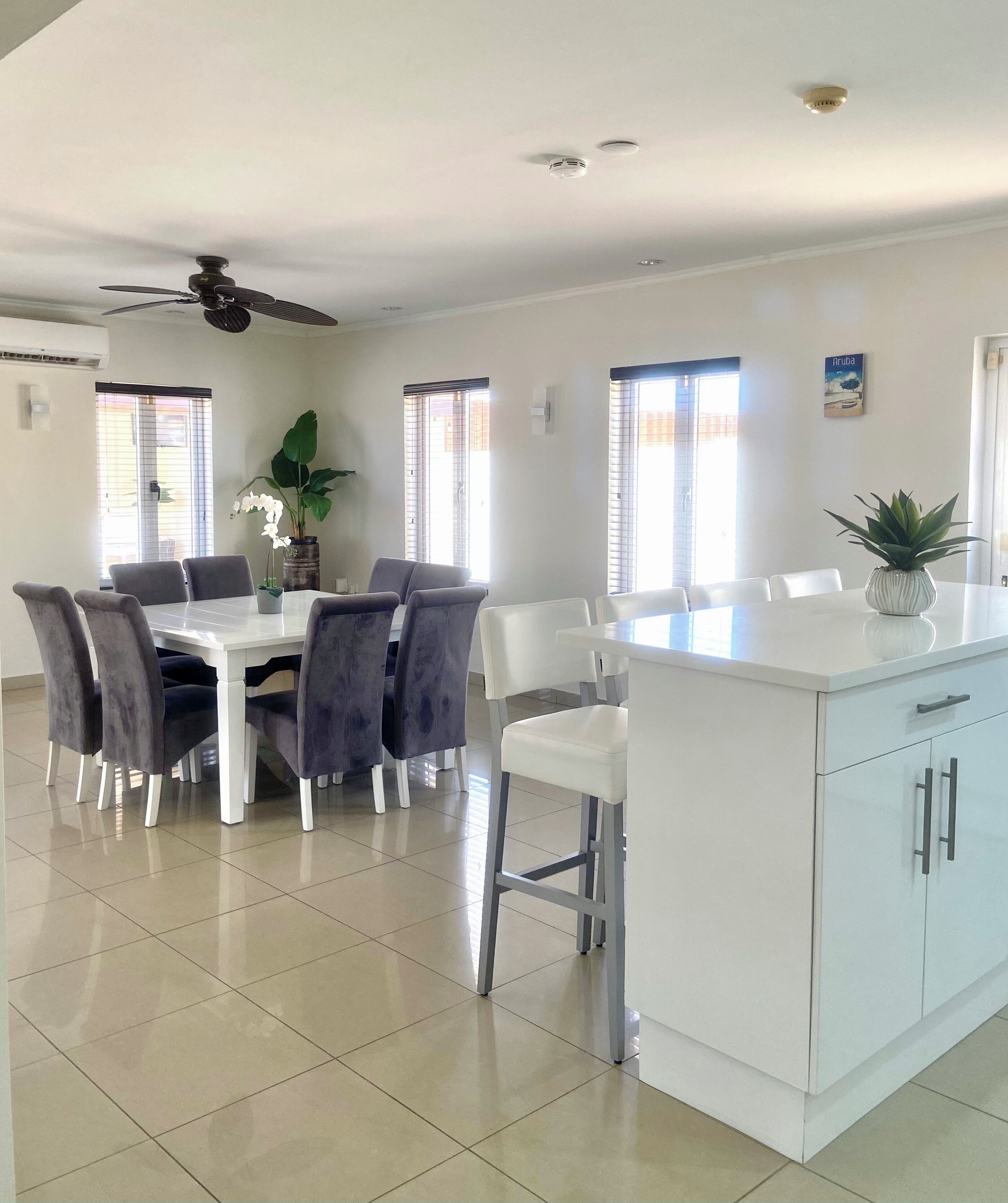 A spacious dining area with a white table and chairs, a ceiling fan, and large windows allowing natural light.