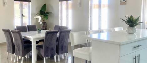 A spacious dining area with a white table and chairs, a ceiling fan, and large windows allowing natural light.