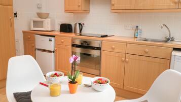 Una cocina moderna con armarios de madera, microondas, hervidor de agua y lavavajillas. Hay una mesa redonda con una planta y cuencos de fruta.