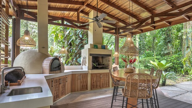 Un área de cocina al aire libre con horno empotrado, una mesa de comedor con sillas y una lámpara colgante.