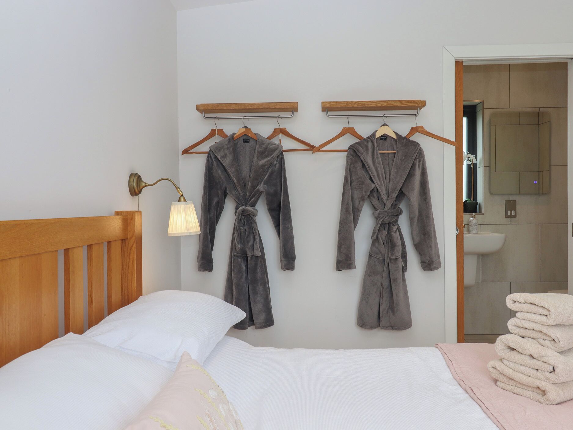 A bedroom with a bed, two robes hanging on a wall, a bedside lamp, and a bathroom visible through an open door.