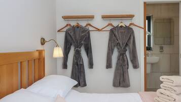 A bedroom with a bed, two robes hanging on a wall, a bedside lamp, and a bathroom visible through an open door.
